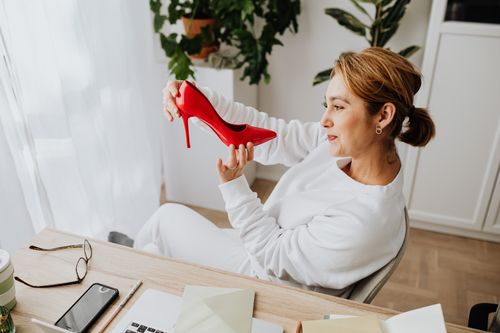 Free Woman in white sweater smiling at red high heel shoe at a desk with plants. Stock Photo