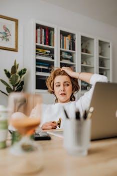 Adult woman working at home office desk with laptop and notepad, looking thoughtful.