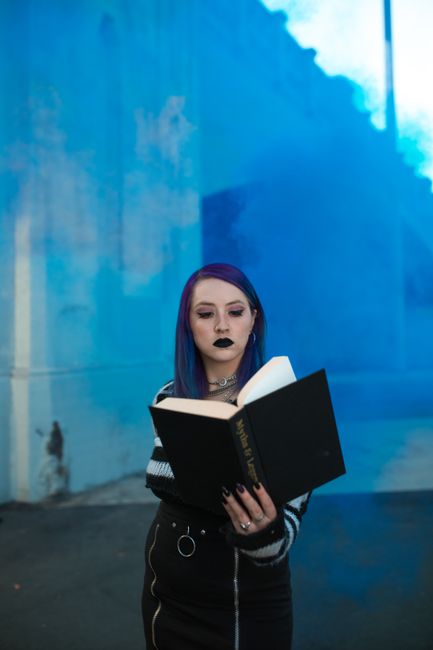 A gothic-styled woman with dark makeup reading a book surrounded by blue smoke outdoors.