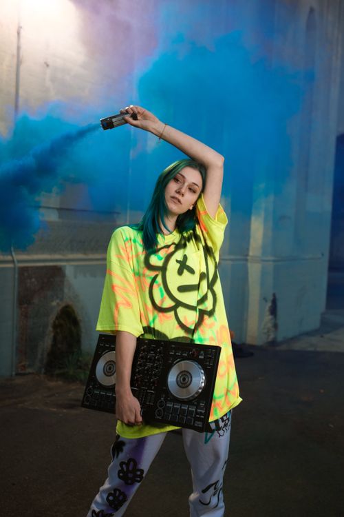 Free DJ in vibrant streetwear holding DJ controller and smoke bomb for a trendy, urban scene. Stock Photo