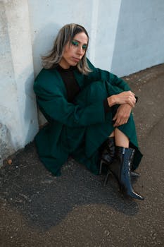 A fashion-forward androgynous adult in a green coat and boots, sitting outdoors with a cool, edgy vibe.