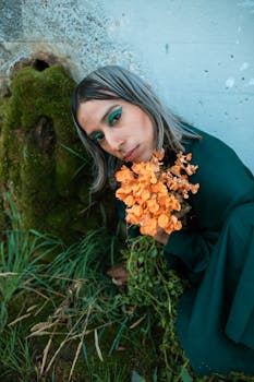 Portrait of a person with vibrant makeup holding orange flowers beside a mossy wall.