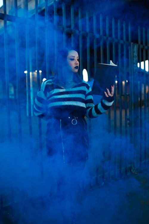 Free A young woman in striped sweater reading a book amidst blue smoke, creating a mysterious vibe. Stock Photo