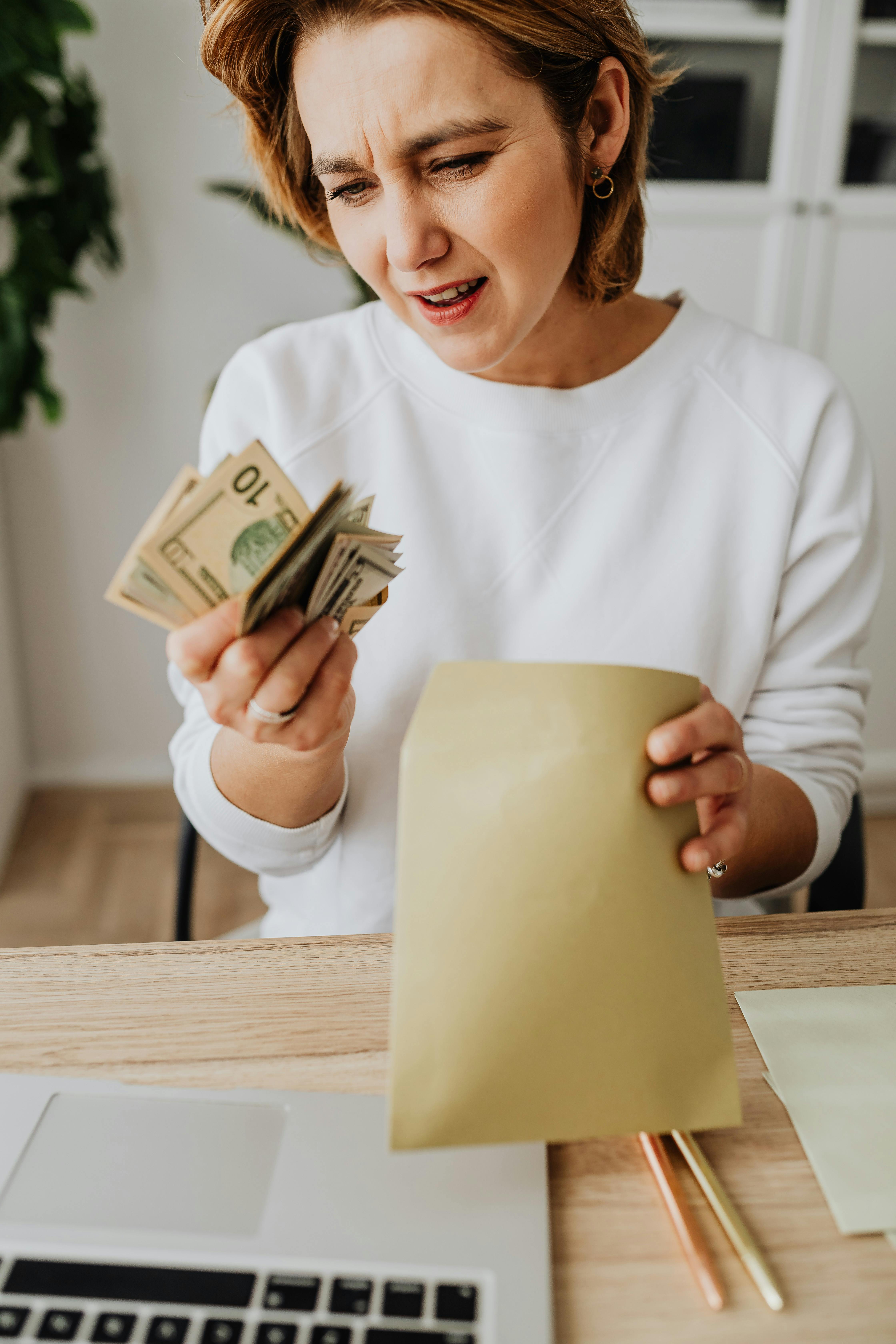 A woman seated at a desk is holding cash and an envelope, looking worried, indoors.