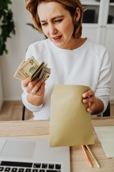 一名妇女坐在办公桌前，手里拿着现金和信封，看起来忧心忡忡