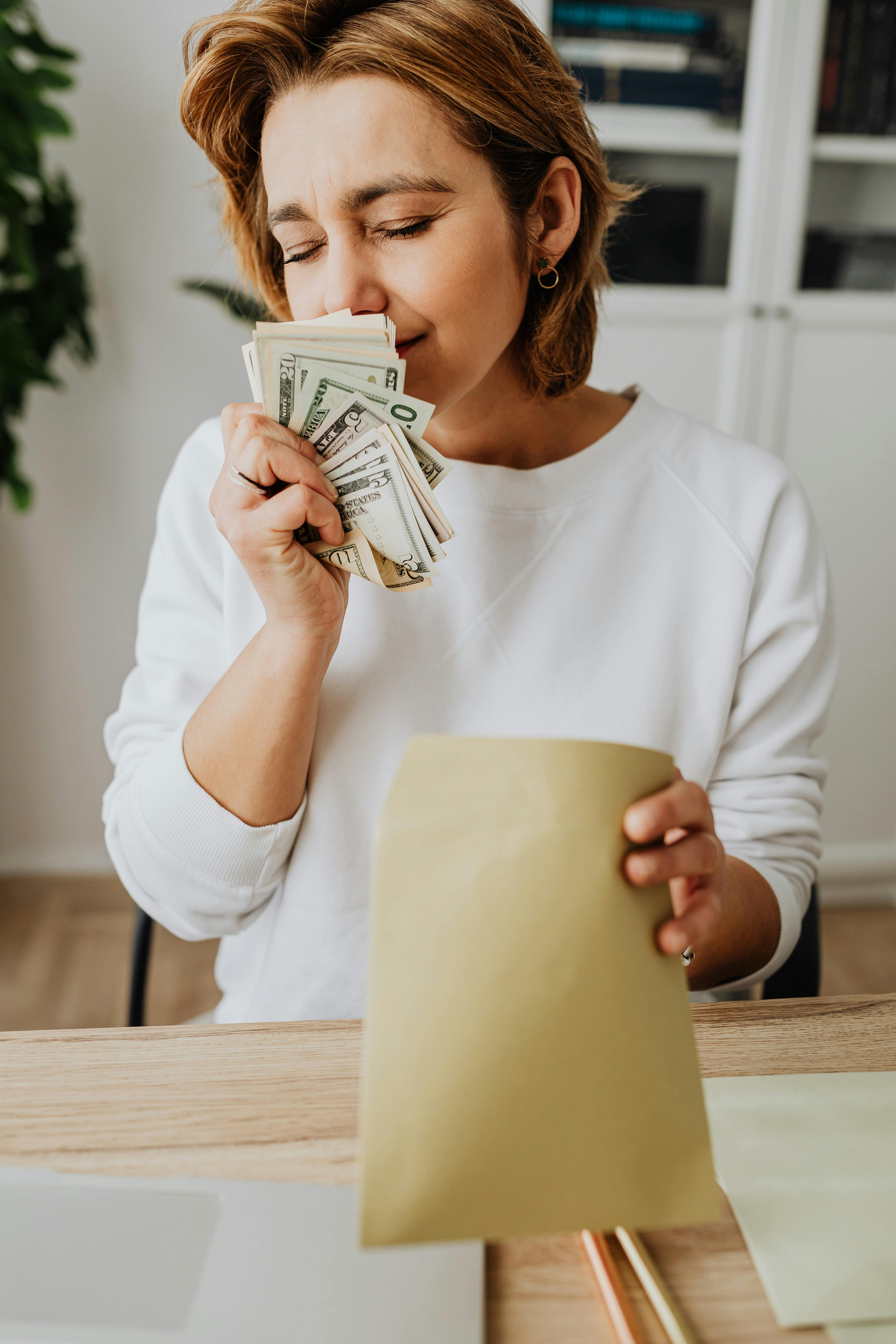 Woman joyfully holding cash and envelope at home desk, savoring her earnings.