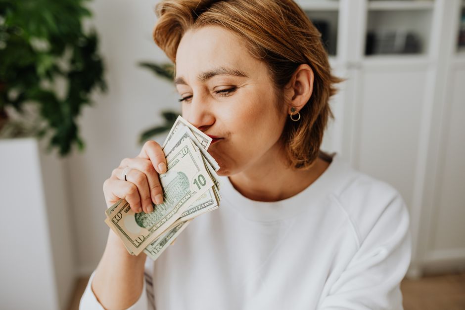 A woman joyfully smelling a handful of US dollar bills indoors.
