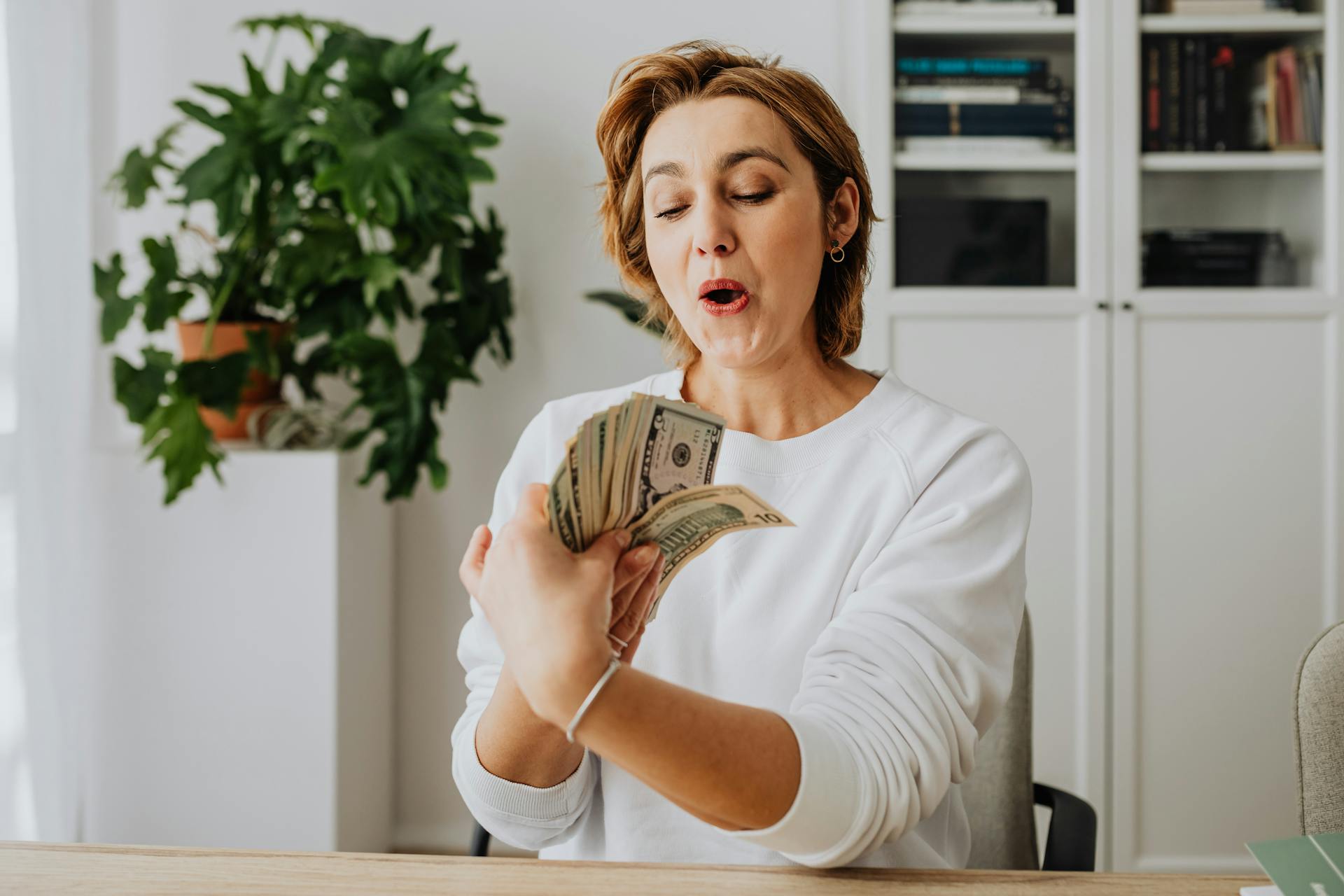 A woman in a white sweater excitedly counts American dollar bills indoors, reflecting joy and prosperity.