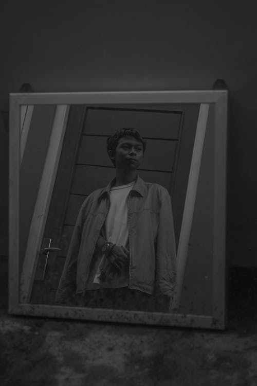 Black and white of serious young Asian male in trendy outfit standing in front of mirror near door and looking away
