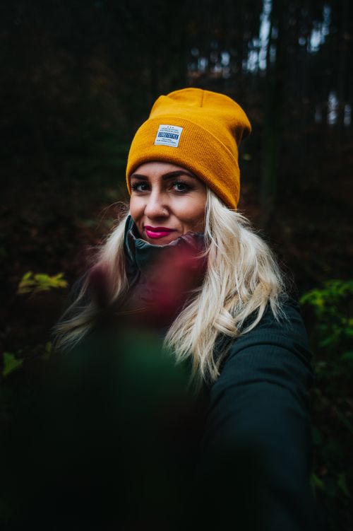 Free Content young lady in warm jacket and hat standing in forest with green trees and shrubs holding plant in hand looking at camera Stock Photo