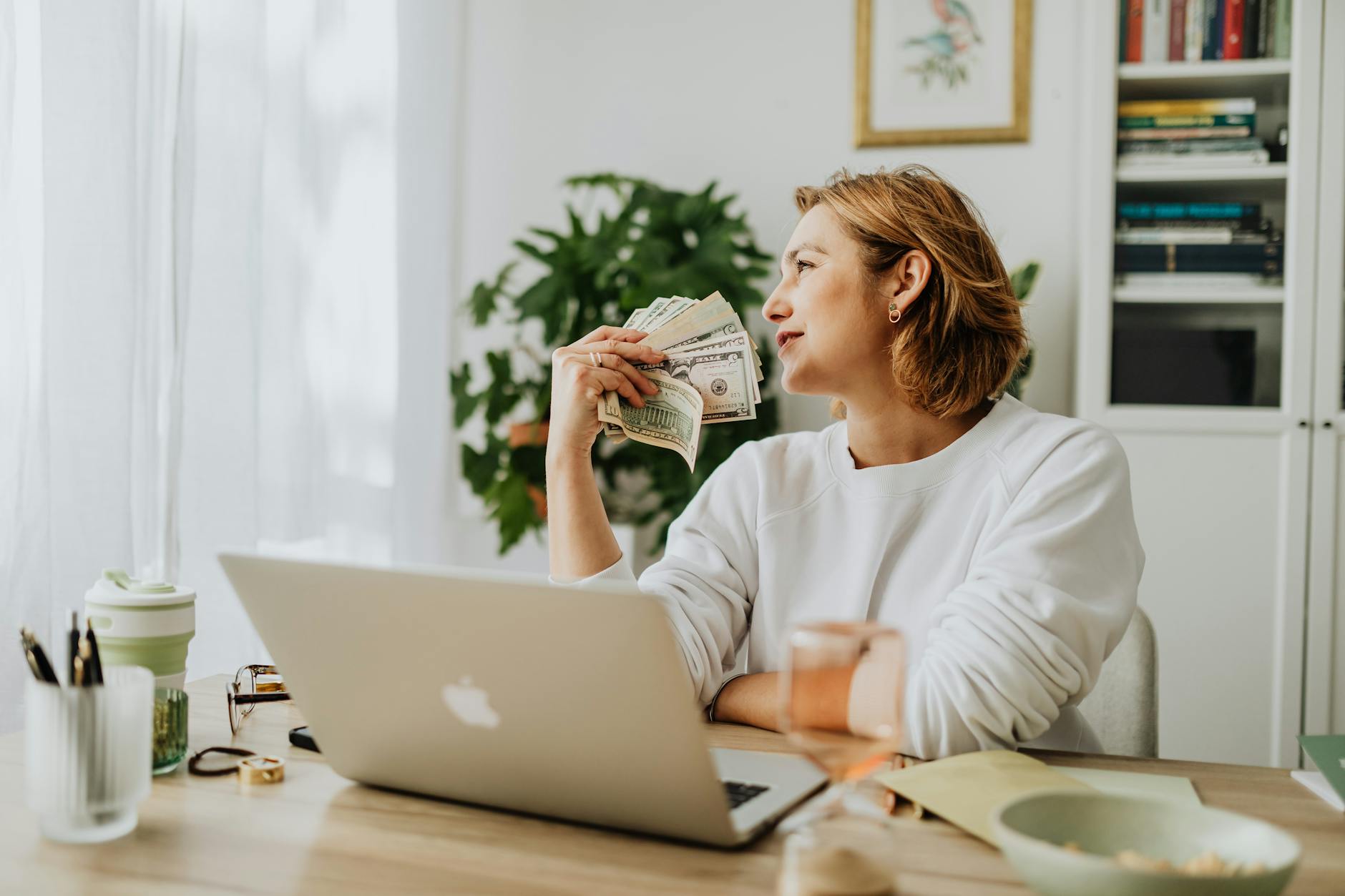 Woman sitting at desk holding cash while working on a laptop in a cozy home office setting.