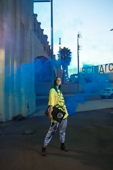 Young adult poses with DJ equipment in vibrant street fashion amidst colorful smoke in an urban setting.