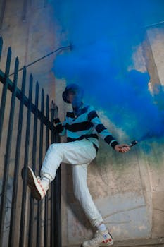 A young adult creating a vibrant scene with a blue smoke bomb against a city backdrop.