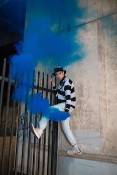 A young adult showcasing urban street style with striking blue smoke against a city backdrop.