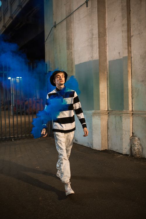 Free Young adult walking through vibrant blue smoke in urban setting, showcasing street style. Stock Photo