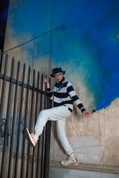 Young man in streetwear leaping by a fence with vibrant blue smoke, urban setting.