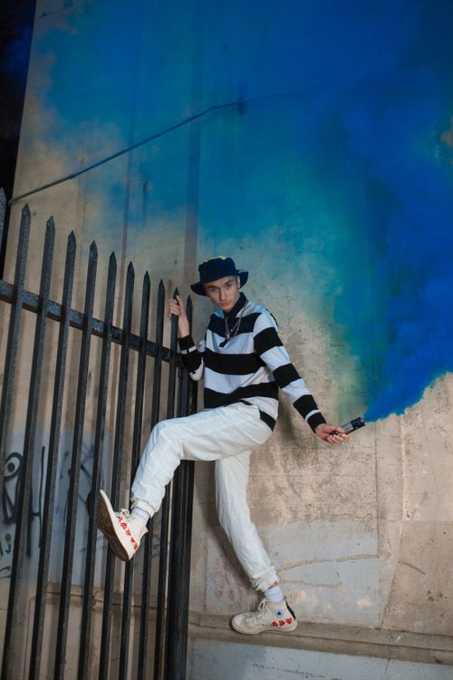 Free Young man in streetwear leaping by a fence with vibrant blue smoke, urban setting. Stock Photo