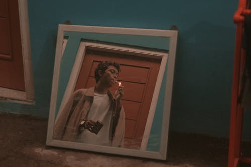 Square mirror placed on floor in room and reflecting young rebellious Asian guy in stylish outfit smoking cigarette near door