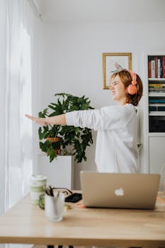Woman stretching in a bright home office wearing headphones, with a laptop on the desk.