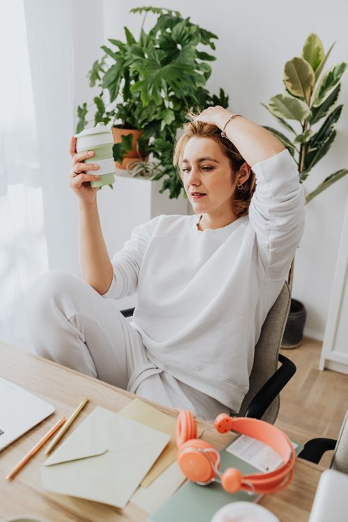 Free Woman taking a break in home office, holding coffee, surrounded by plants. Stock Photo