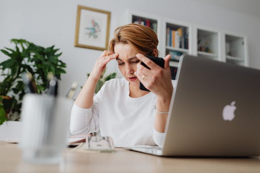A stressed woman holds her phone, working at a desk with laptop and papers.