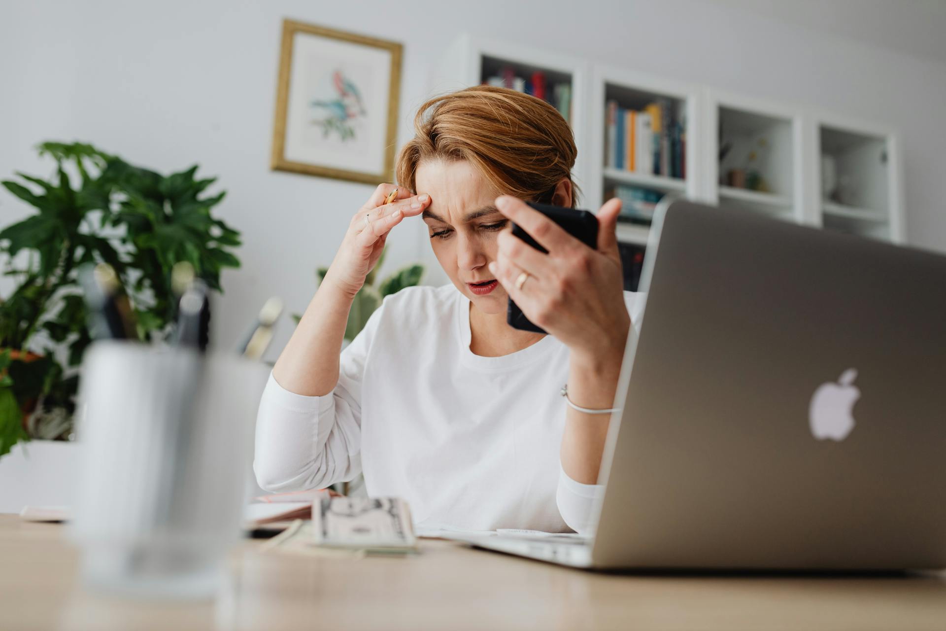 A stressed woman holds her phone, working at a desk with laptop and papers.