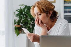 A woman reviews financial documents at a home desk with a laptop and cash visible.