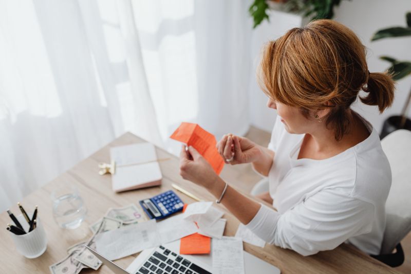A woman at a sunlit desk holding a paper slip, with more receipts, two calculators and a pen cup on the table.