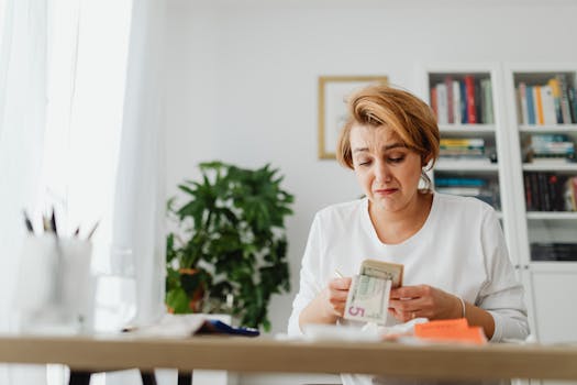 A woman concernedly counts money at her desk in a bright room.
