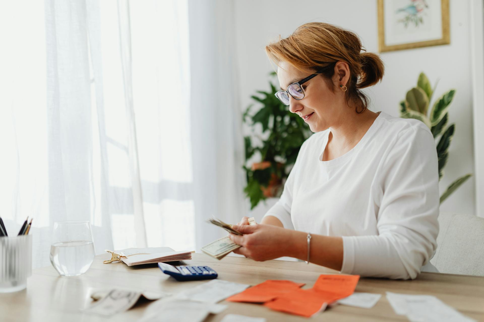 Woman counting money at home desk with papers and calculator, emphasizing financial management.