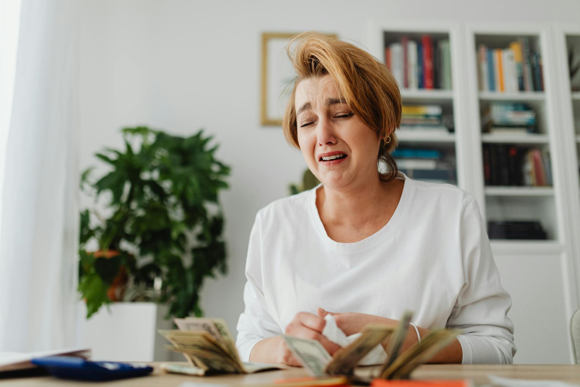 A distressed woman counts cash at a desk with a pained expression, highlighting financial strain.