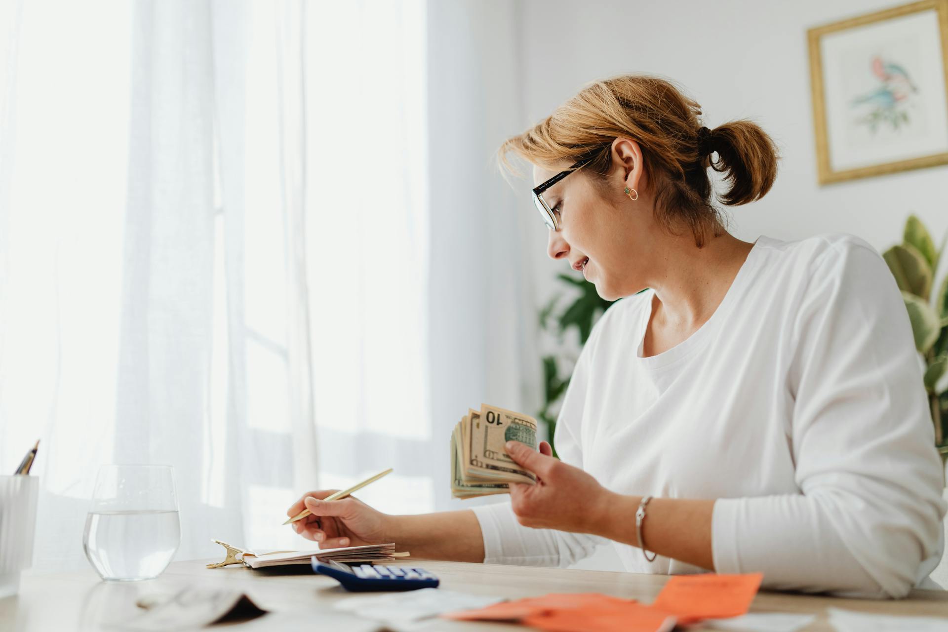 Businesswoman calculating finances with cash, calculator, and notebook at a bright desk.