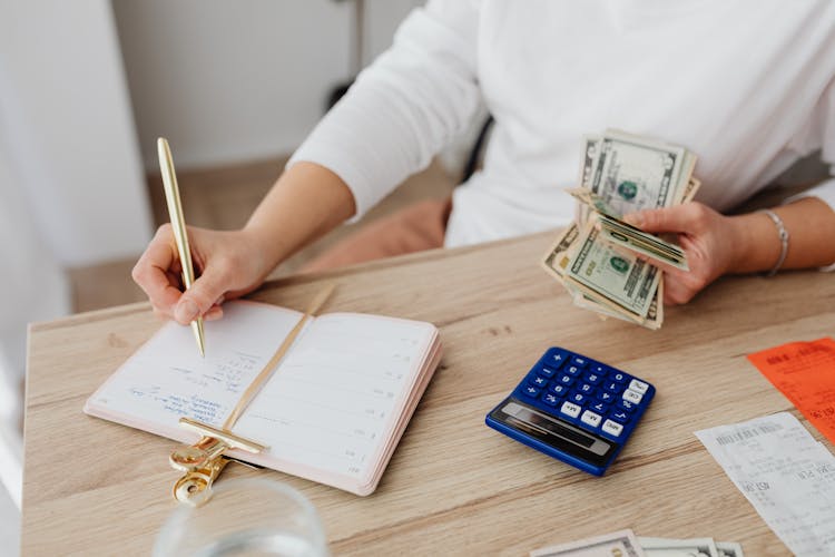 Person Writing On A Notebook While Holding Money 