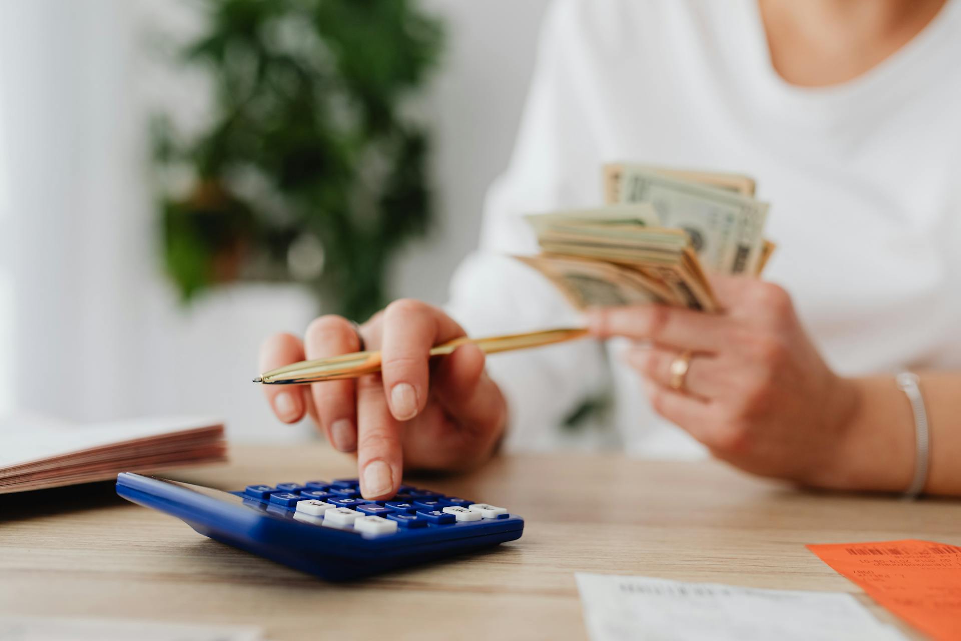 A person sits at a desk calculating finances using a calculator and holding cash.