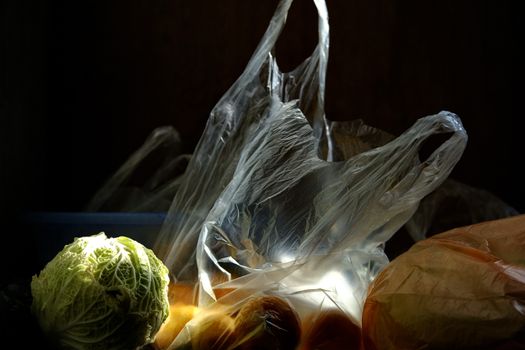 Still life of a cabbage and plastic bags with fruits illuminated in dim light, highlighting texture and shadow contrast.