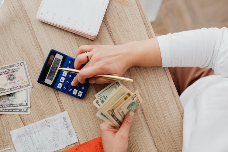 Person Using A Blue Calculator While Holding Dollar Bills 