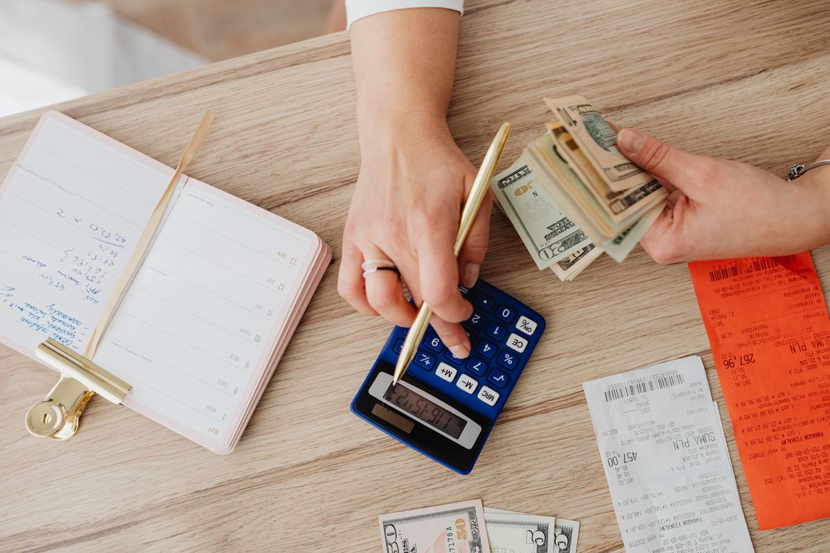 Two hands counting dollar bills over a table that holds a calculator and a stack of bills.