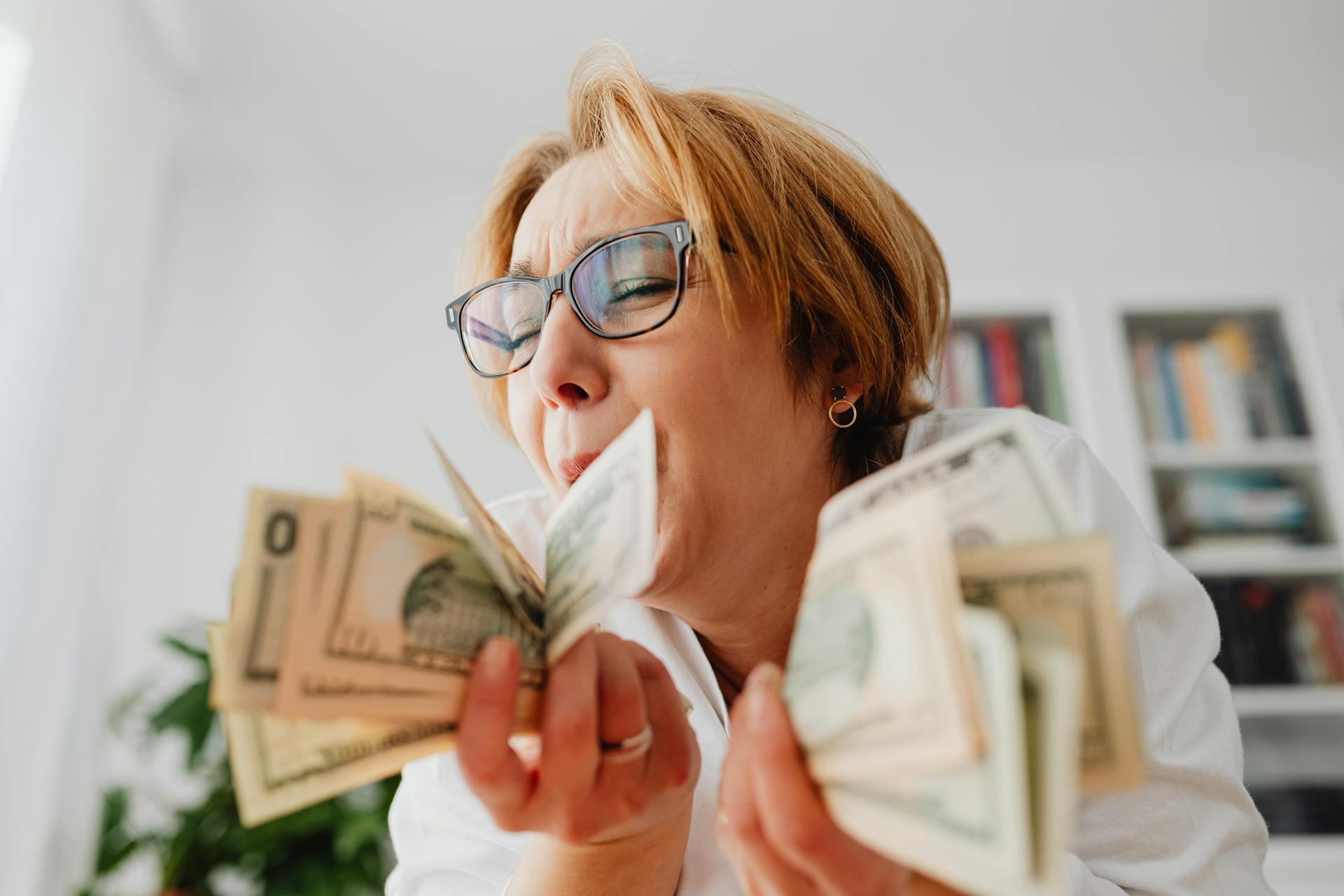 A happy woman with eyeglasses cherishing US dollar bills indoors, symbolizing wealth and financial success.