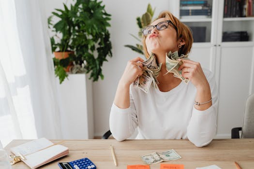 A happy woman holding dollar bills, sitting at a desk with plants and office supplies.
