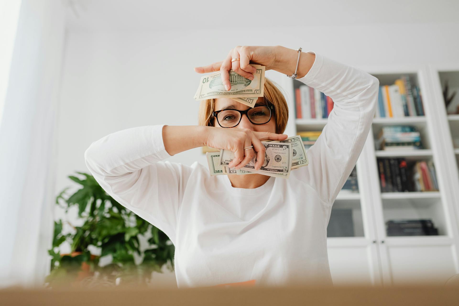 Smiling woman indoors, playfully holding cash in hands, expressing joy.