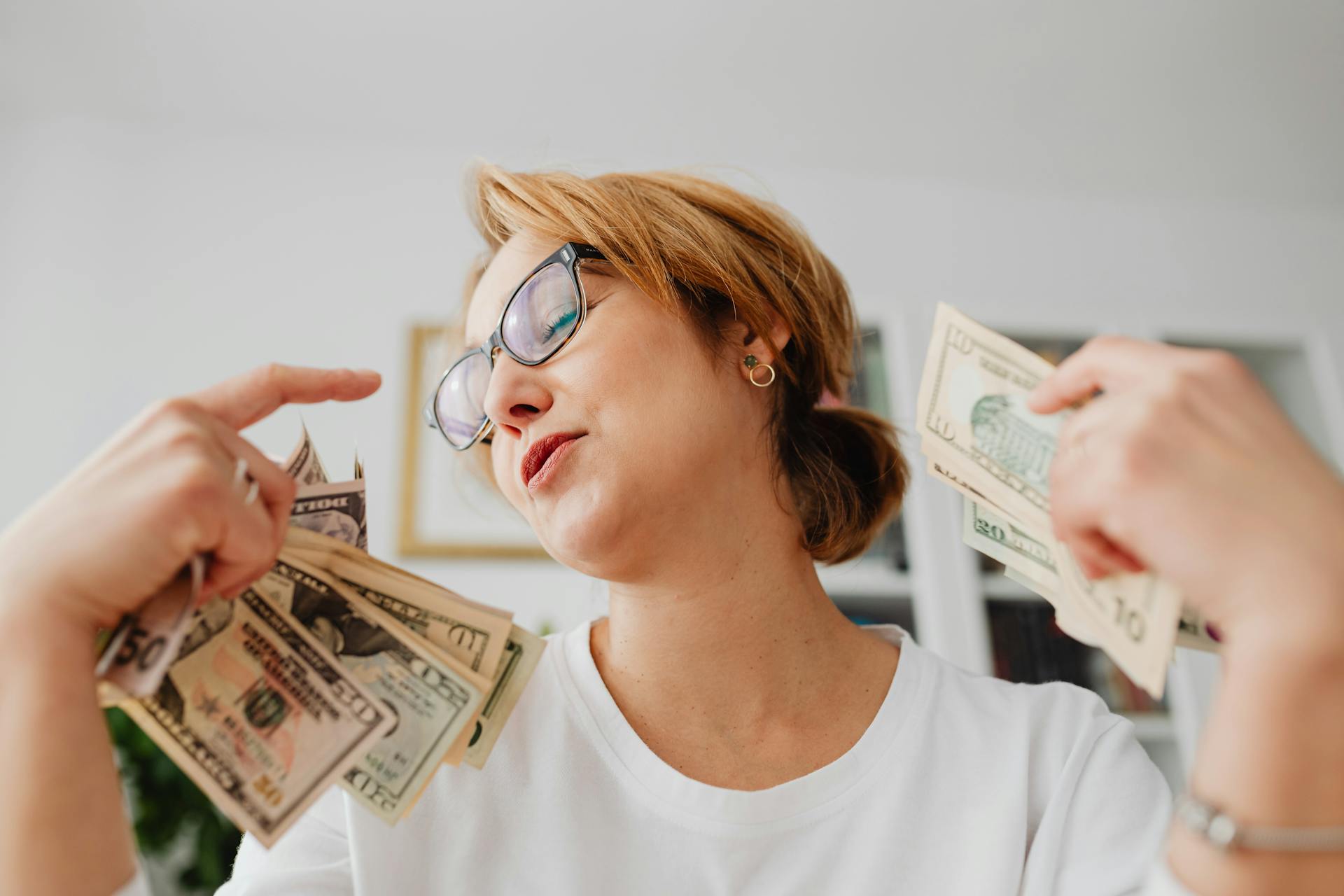 Confident woman wearing glasses and holding US dollar bills in both hands indoors.