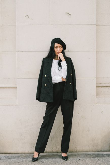 Stylish woman in black blazer and pants, posing against a wall outdoors.