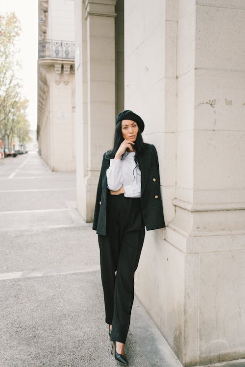 бесплатная Женщина в черном пиджаке и черных штанах стоит у белой бетонной стены Стоковое фото