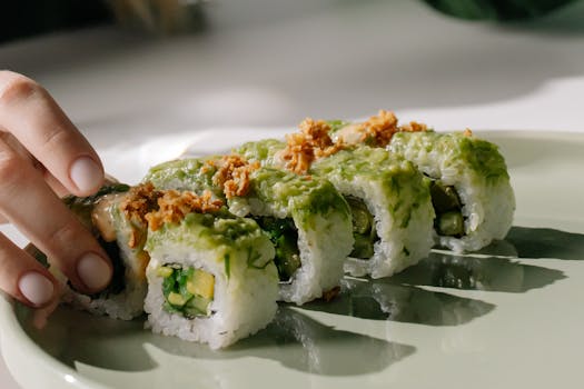 A hand reaches for a sushi roll topped with avocado and fried onions on a green plate.