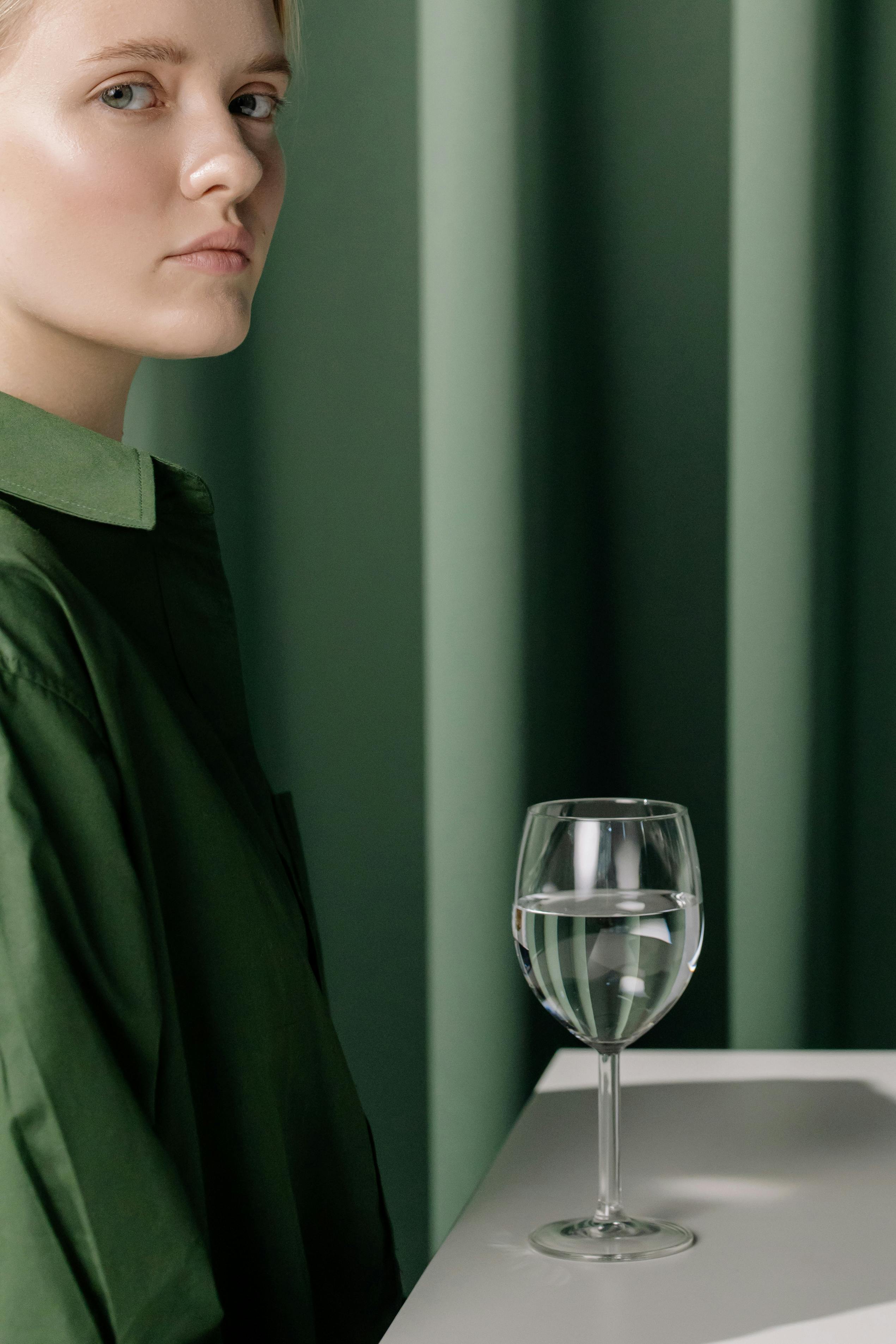A woman in a green shirt with a serious expression sits beside a glass of water indoors.