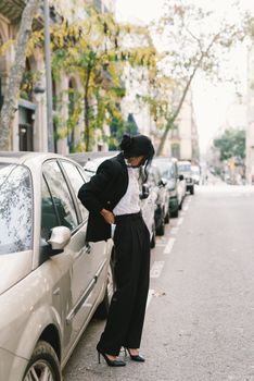 一位穿着黑色外套和高跟鞋的时尚女子，站在绿树成荫的街道旁，停在一辆汽车边