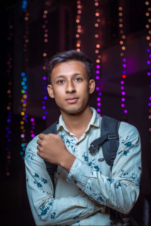 Free Portrait of a young man with a backpack standing against a backdrop of colorful string lights at night. Stock Photo