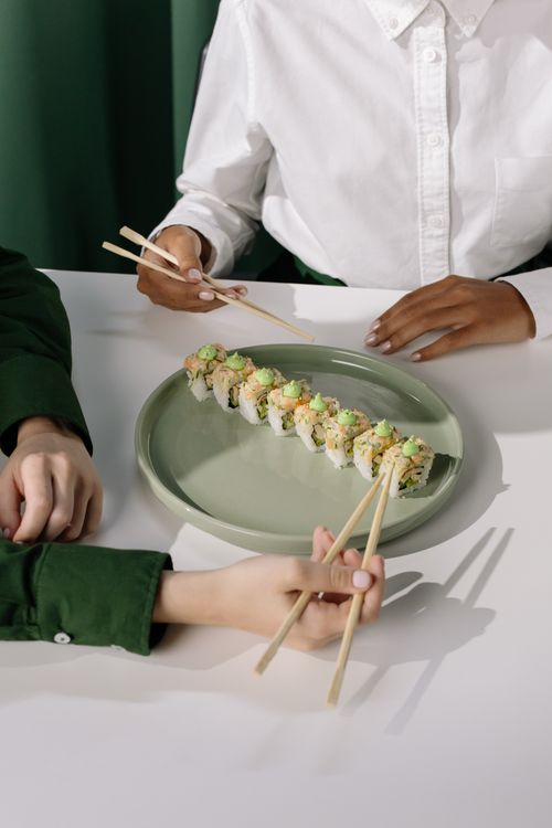 Free Two individuals using chopsticks to pick sushi rolls from a green plate, creating a vibrant dining scene. Stock Photo