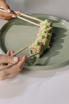 Hands delicately using chopsticks to prepare a sushi roll on a ceramic plate, showcasing traditional Japanese cuisine.