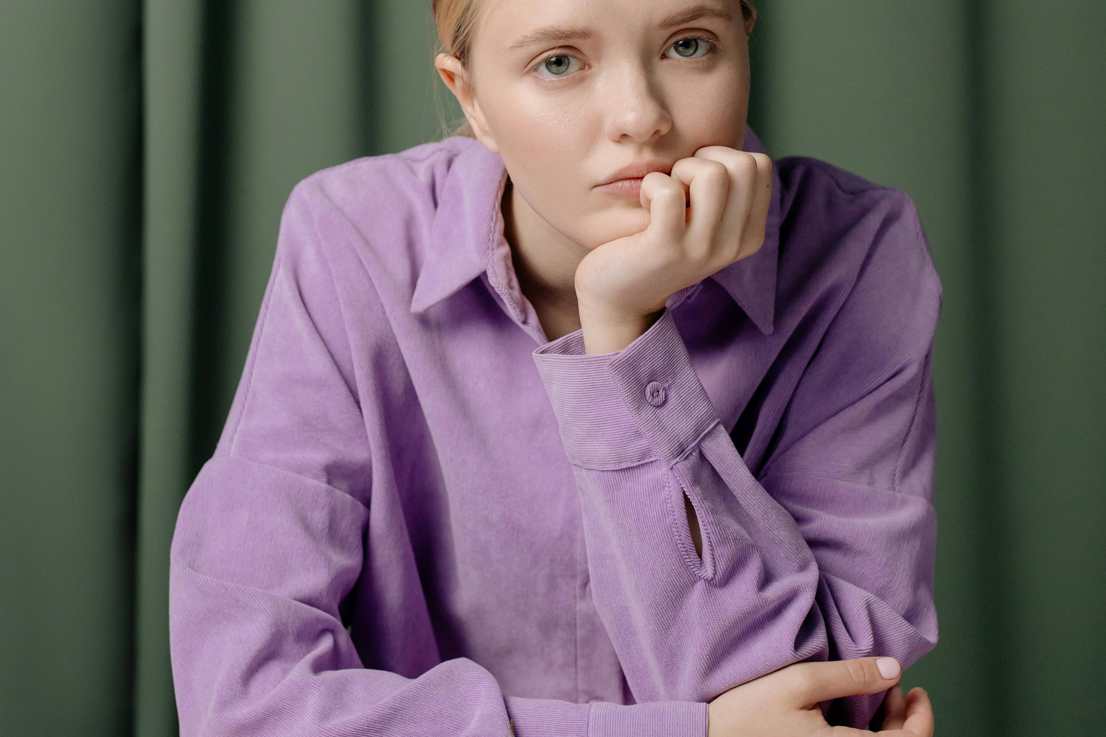 Portrait of a young woman in a purple shirt with hand on chin, looking thoughtful.
